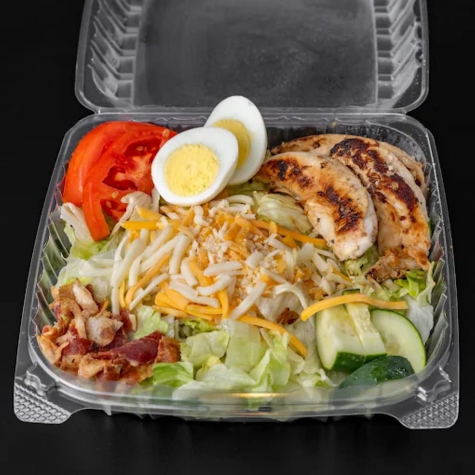Grilled chicken salad with tomatoes, hard-boiled eggs, cheese, bacon, and cucumbers in a plastic container.